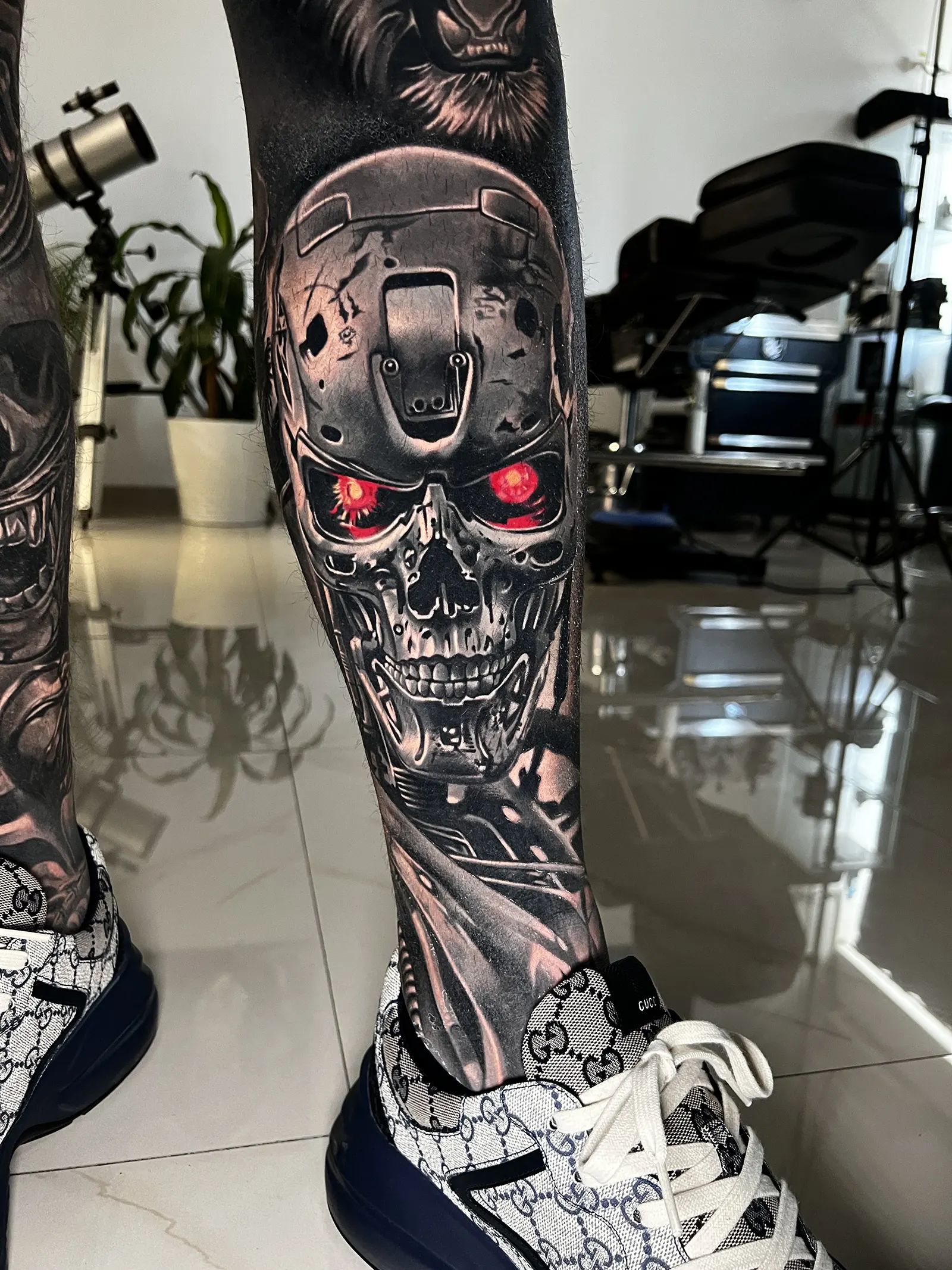Tatuaj realist alb-negru cu craniu robotic și ochi roșii, realizat pe gambă