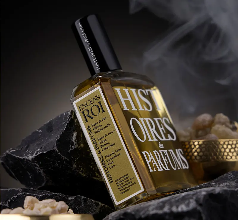 Sticla parfumului ENCENS ROI așezată pe roci negre, în decor cu fum